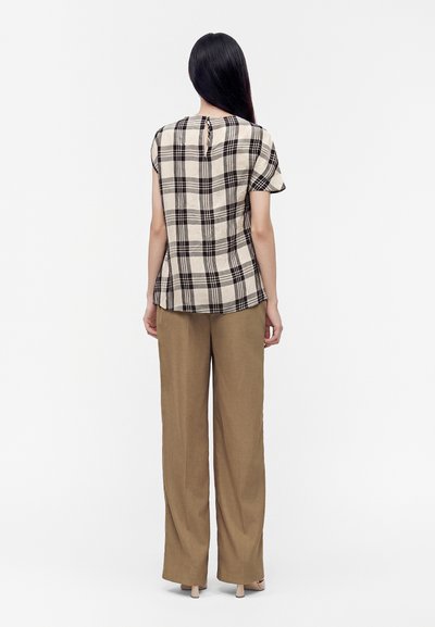 Femme aux longs cheveux noirs portant un haut à manches courtes à carreaux noir et blanc et un pantalon marron ample, debout de dos.