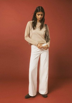 Beżowy sweter, białe szerokie dżinsy z ćwiekami po bokach oraz ciemne buty. Tło jest jednolite, czerwone.