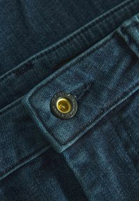 Temno modri denim z teksturirano površino; vključuje kovinsko gumbo z zlatim srednjim delom in temnim zunanjim robom ter šivane roba.