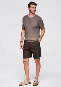 Camicia a maniche corte marrone a coste con tre bottoni, abbinata a pantaloni corti scuri e sandali beige, caratterizzata da un tessuto testurizzato e una vestibilità rilassata.