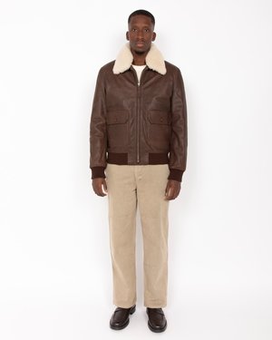 Veste en cuir marron avec un col en fausse fourrure crème, des poches avant et des poignets côtelés, associée à un pantalon en velours côtelé beige et des chaussures marron.