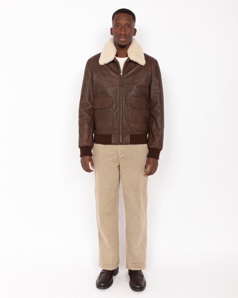 Chaqueta de cuero marrón con cuello de borreguito sintético crema, bolsillos frontales y puños acanalados, combinada con pantalones de pana beige y zapatos marrones.