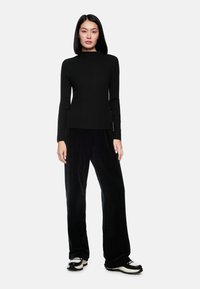 Schwarz gestreiftes Langarmoberteil kombiniert mit schwarzen weiten Cordhosen. Das Modell trägt schwarze und weiße Sneakers. Einfaches, körpernahes Design.