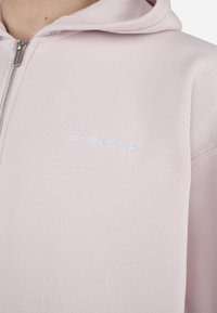 Sudadera con cremallera de color rosa claro, con un tejido texturizado, que presenta un logotipo bordado blanco en el pecho y una capucha con cordón ajustable.