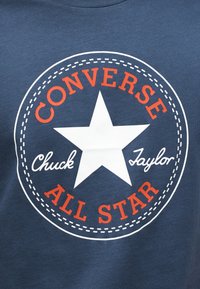 Μπλε ύφασμα με ένα λευκό αστέρι στο κέντρο, περιβαλλόμενο από πορτοκαλί κείμε νο «CONVERSE ALL STAR» και λευκή καμπύλη γραφή «Chuck Taylor».