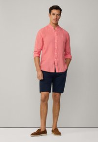 Licht koraalkleurig linnen shirt met een bandkraag, knoopsluiting en opgerolde mouwen, gecombineerd met marineblauwe shorts en bruine instappers.