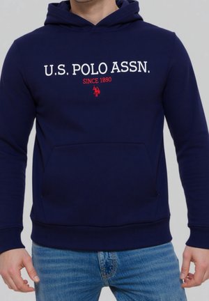 Mężczyzna w granatowej bluzie z kapturem z napisem "U.S. POLO ASSN. SINCE 1890" i czerwonym logo gracza polo, w połączeniu z niebieskimi dżinsami.