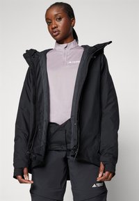 Sort zip-up jakke med hætte over en lys lilla top; har en glat tekstur og funktionelt design. Logo synligt på toppen og bukserne.