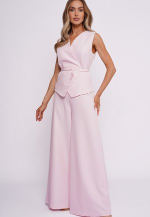 Kobieta z długimi falowanymi włosami, ubrana w bezrękawnik w kolorze jasnoróżowym z kopertowym dekoltem oraz szerokie spodnie w tym samym kolorze, stojąca na tle gładkiego tła.