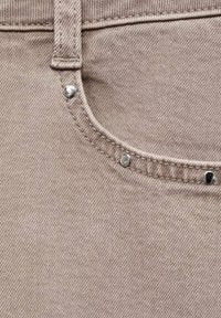 Tela de mezclilla beige con una textura sutil, que presenta detalles de tachuelas metálicas en la cintura y costuras elaboradas.