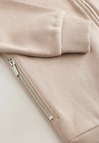 Felpa rosa chiaro con una superficie liscia, dettagli con tasche con zip e polsini a costine. Presenta un design semplice e non decorato.