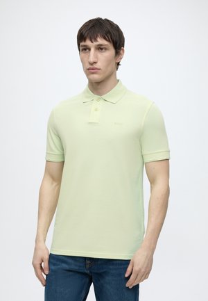 Mand iført lysegrøn kortærmet poloshirt og blå jeans, stående mod en ensfarvet hvid baggrund.