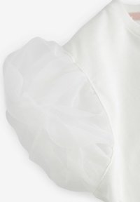 Haut blanc en coton doux et texturé, avec des manches en tulle transparent et volantes, ajoutant du volume et une touche délicate.