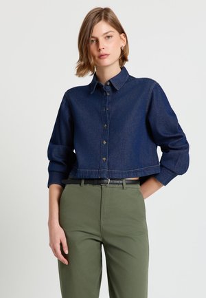 Jeune femme portant une chemise en jean bleu foncé raccourcie avec les manches retroussées et un pantalon vert olive avec une ceinture noire, sur un fond uni.