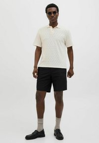 Gaišs krēmveida polo krekls ar rakstu un mīkstu apkakli, apvienots ar melniem šortiem un tumšām kurpēm. Modelis valkā tumšas apaļas saulesbrilles.