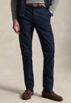 Mann trägt marineblaue Hose, braunen Gürtel, grauen Pullover und braune Loafers, steht mit einer Hand in der Tasche vor neutralem Hintergrund.