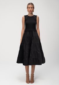 Czarna sukienka z ryfowanym korpusem, bez rękawów i warstwowym dołem. Zawiera guziki na ramionach oraz rozszerzaną, teksturowaną dolną część.