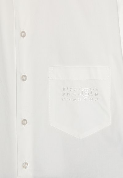 Vit skjorta med knappar och ficka, prydd med diskret broderi av siffror och en cirkel. Slät textur och standardkragsdesign.