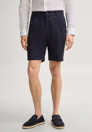 Mann trägt marineblaue Shorts, weißes langärmeliges Hemd und dunkle Slipper, steht vor einem schlichten Hintergrund.
