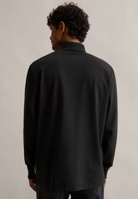 Μαύρο μπλουζάκι με μακριά μανίκια, φτιαγμένο από λείο ύφασμα. Διαθέτει σταντ γιακά, χαλαρή εφαρμογή και μινιμαλιστικό σχέδιο. Χωρίς ορατά μοτίβα ή αξεσουάρ.