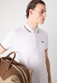 Hvit polo-skjorte med svart og beige stripete krage, med en liten svart logo. Modellen holder en brun lærveske med doble håndtak.
