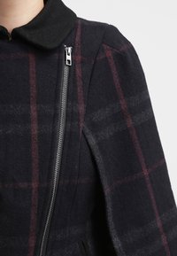 Manteau en laine à carreaux noir et bordeaux, doté d'une fermeture éclair, d'un col arrondi et de poches latérales avec une texture douce.