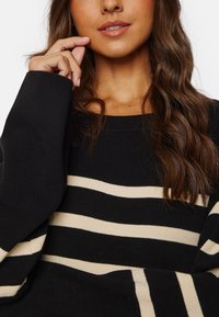 Maglione nero con ampi strisce beige, caratterizzato da un colletto e polsini a coste. Texture morbida con una vestibilità ampia e oversize.