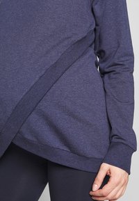 Camiseta de manga larga azul marino con dobladillo curvado y puños acanalados. Textura suave, que presenta un diseño en ángulo con una apariencia de capa.