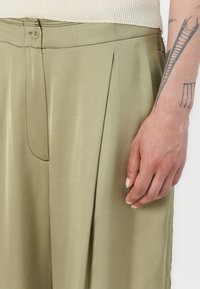 Olive green, pleated trousers made of soft fabric, featuring a front button closure and side pockets. Close-up of hand resting on the fabric.