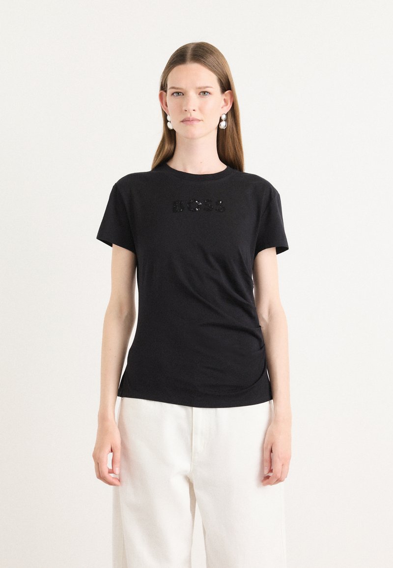 T-shirt in cotone nero con maniche corte, caratterizzato da un logo 'BOSS' textured con paillettes sul davanti. Abbinato a pantaloni chiari.