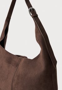 Sac épaule en suède brun avec une forme incurvée, présentant une boucle en métal argenté sur la sangle et des détails de couture discrets le long des bords.