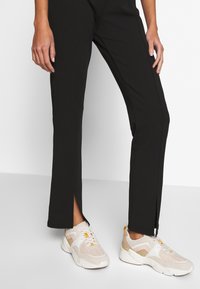 Černé kalhoty s rovným střihem a bočními rozparky, doplněné béžovobílými teniskami s síťovaným svrškem a strukturovanou podrážkou.