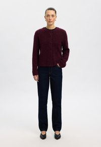 Vinröd stickad kofta med rund halsringning och knäppning framtill, kombinerad med mörkblå jeans med rakt ben och svarta skor med spetsig tå.