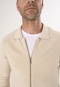 Beige geribde cardigan met een rits, lichtgewicht brei-textuur en een kraag. Draagbaar over een wit t-shirt, die een eenvoudig ontwerp laat zien.