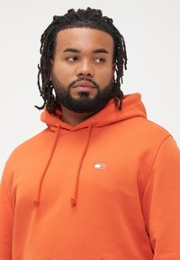 Uomo con barba e dreadlocks che indossa una felpa arancione con il logo "Tommy Jeans" mentre guarda a sinistra su uno sfondo neutro.