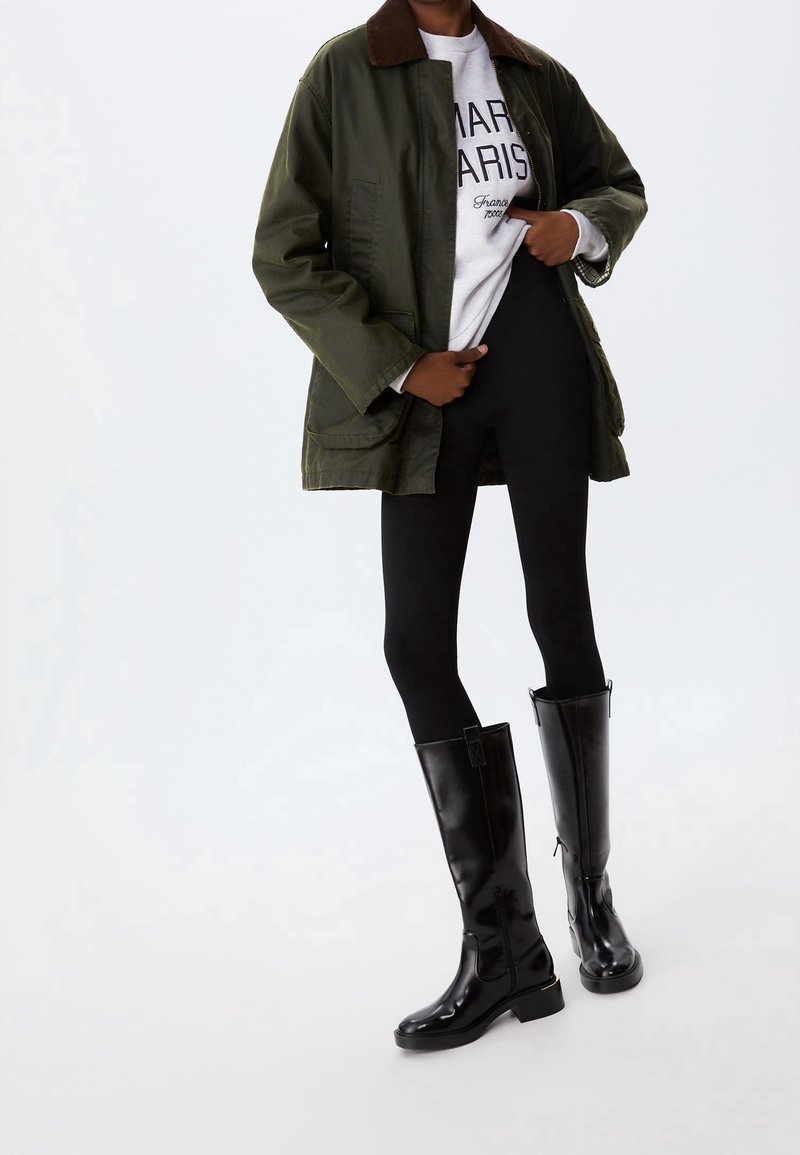 Personne portant une veste vert olive, un sweat-shirt blanc avec un texte, des leggings noirs et des bottes en cuir noires montant jusqu'aux genoux, debout sur un fond uni.