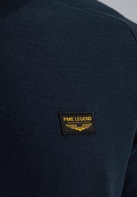 Navyblauwe gebreide trui met een opvallend rechthoekig zwart label met gouden tekst en vleugeldesign, dat het merk PME Legend aangeeft.
