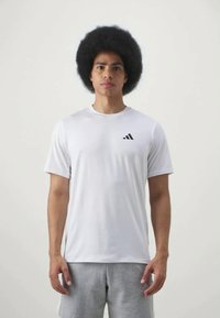 Weißes Sportshirt aus leichtem Stoff, mit einem schwarzen Adidas-Logo auf der linken Brust, kombiniert mit grauen Shorts.