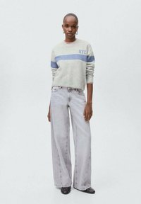 Šedý svetr s modrým pruhem a textem "NYC", spárovaný s světle šedými širokými džiny. Modelka má na sobě černé slip-on boty se hroty.