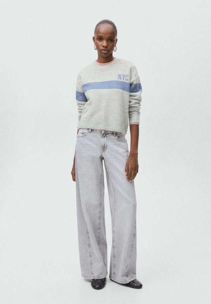 Šedý svetr s modrým pruhem a textem "NYC", spárovaný s světle šedými širokými džiny. Modelka má na sobě černé slip-on boty se hroty.