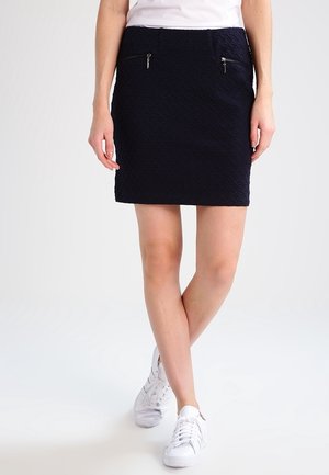 Femme portant une mini-jupe noire texturée avec deux poches zippées à l'avant, un haut blanc et des baskets blanches, debout avec les jambes croisées.
