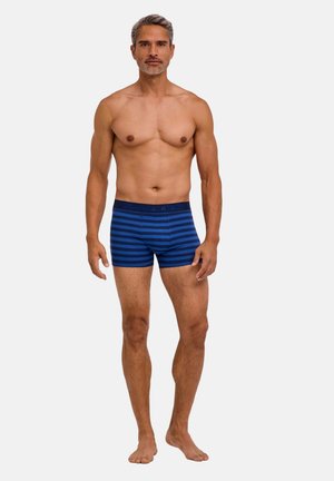 Fit voksen mand står barfodet iført blå og marineblå stribede boxershorts mod en ensfarvet lys baggrund.