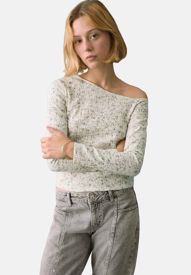 Vapaa-olkapää neulepaita vaaleanharmaana, jossa on teksturoitu kuvio, yhdistettynä harmaisiin farkkuihin, joissa on niittikoristeita ja rento istuvuus.