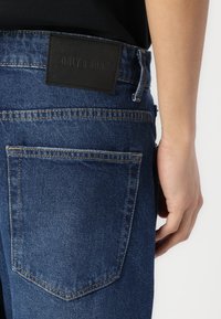 Denim hlače v svetlo do srednje modri barvi z teksturiranim zaključkom. Značilnosti vključujejo črno usnjeno etiketo na pasu in standardne zadnje žepke.