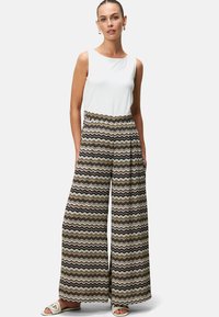 Un pantalon large à motifs dans un design ondulé marron, noir et blanc, associé à un haut sans manches ajusté blanc et des sandales légères.