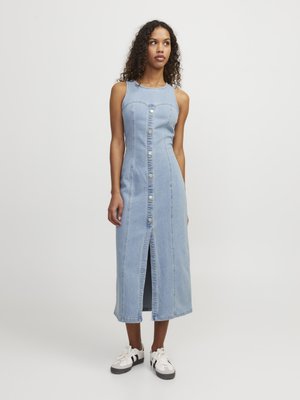 Vrouw draagt een lichtblauwe denim midi-jurk zonder mouwen, met knopen aan de voorkant, een voorsplit en witte sneakers, staand tegen een effen achtergrond.