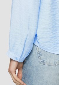 Chemise à manches longues bleu clair, texturée, avec un poignet à boutons, portée avec un jean en denim clair, présentant une coupe décontractée et un tissu doux.