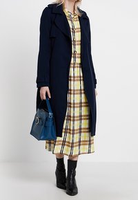 Marineblauer Trenchcoat über einem gelben Karo-Kleid mit braunen Akzenten, kombiniert mit einer blauen Handtasche und schwarzen Stiefeletten. Insgesamt glatte Texturen.