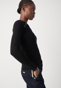 Černý pletený svetr s přiléhavým střihem, který má dekorativní knoflíčky na rameni. Nosený s tmavými denimovými kalhotami se pruhovanými akcenty.