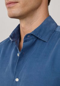 Camicia di velluto a coste blu con colletto button-down, dotata di bottoni chiari e superficie testurizzata. Inquadratura ravvicinata su colletto e scollatura.